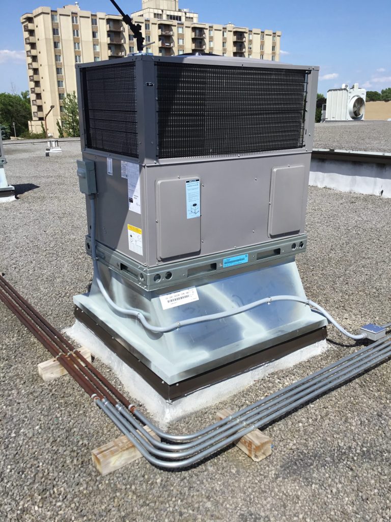 Commercial HVAC unit installed on a flat rooftop, with visible piping and a multi-story building in the background, illustrating Mechanical Heating & Cooling's installation services.