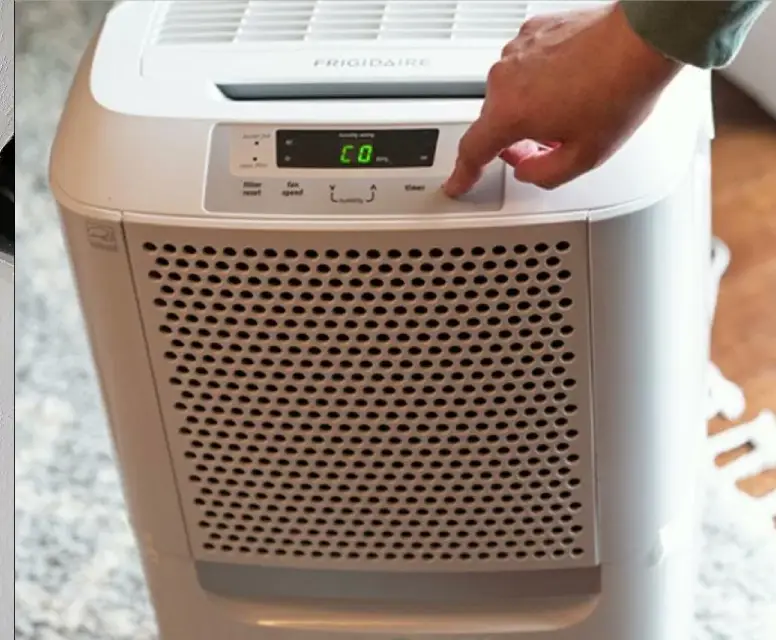 Hand adjusting settings on a Frigidaire dehumidifier, displaying control panel with digital readout, in a home environment.
