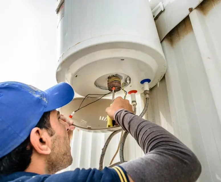 A technician in a blue cap and grey long-sleeved shirt uses a screwdriver to repair the internal electrical components located at the bottom of a white, wall-mounted electric water heater.